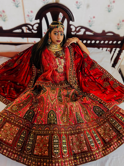 A woman dressed in an ornate red traditional lehenga featuring heavy gold embroidery, mirror work, and intricate motifs, paired with elaborate kundan jewelry and a matching dupatta, posing gracefully on a carved wooden bed; showcasing luxurious Indian bridal fashion craftsmanship.
