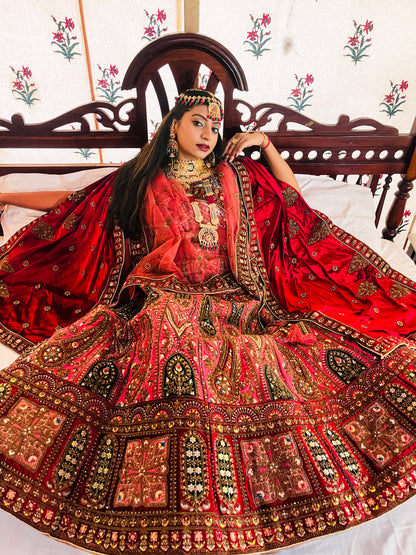 A woman wearing a traditional red bridal lehenga with intricate gold embroidery, heavy kundan jewelry, and a matching dupatta, posing elegantly on a carved wooden bed; showcasing detailed handwork and luxurious Indian ethnic fashion.