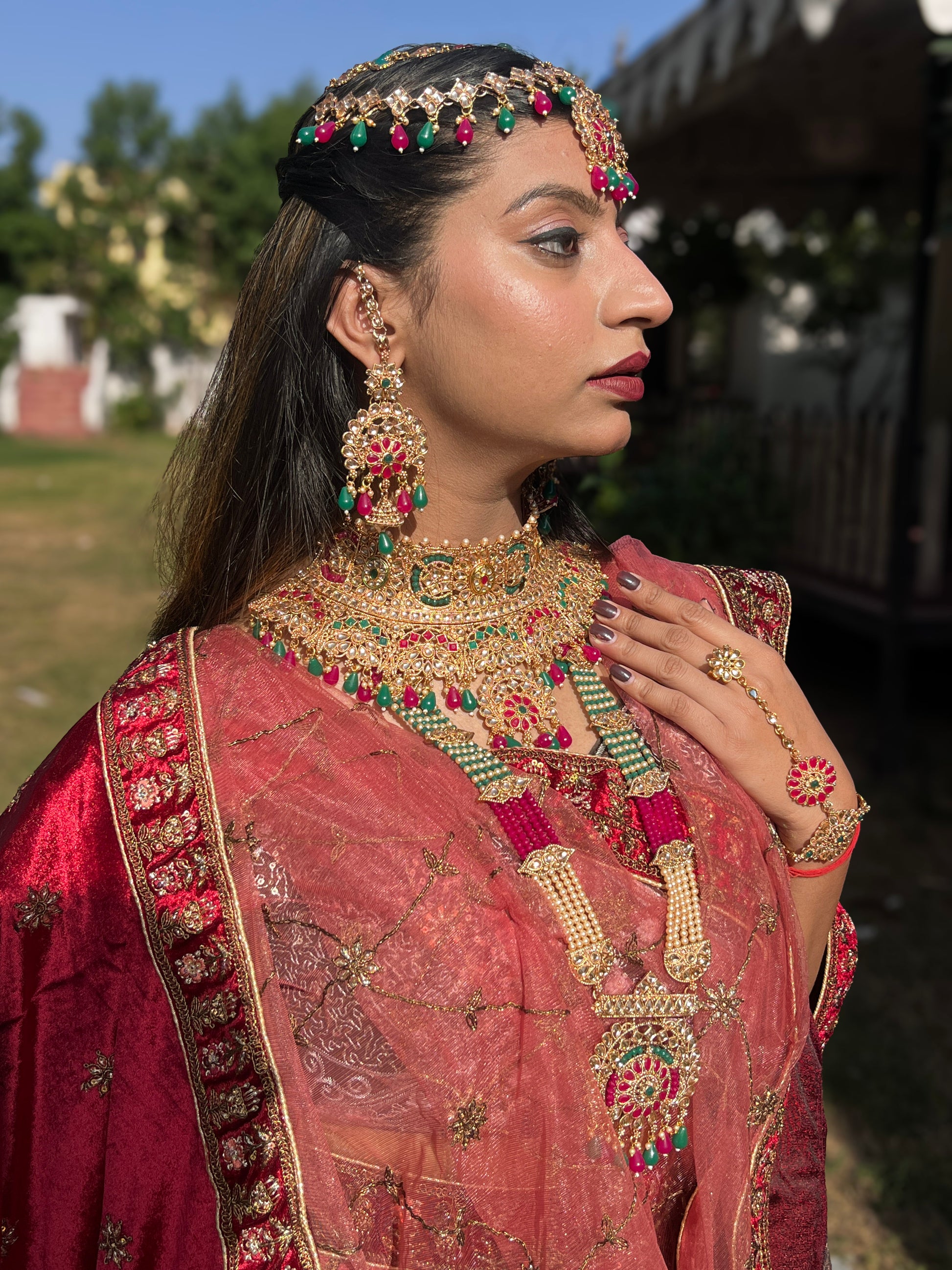 A woman wearing a traditional Indian bridal ensemble from Amai Jaipur, adorned in an embroidered red saree with intricate gold detailing and heavy Kundan jewelry including a maang tikka, necklace, earrings, hand harness, and layered haar, captured outdoors in natural sunlight.