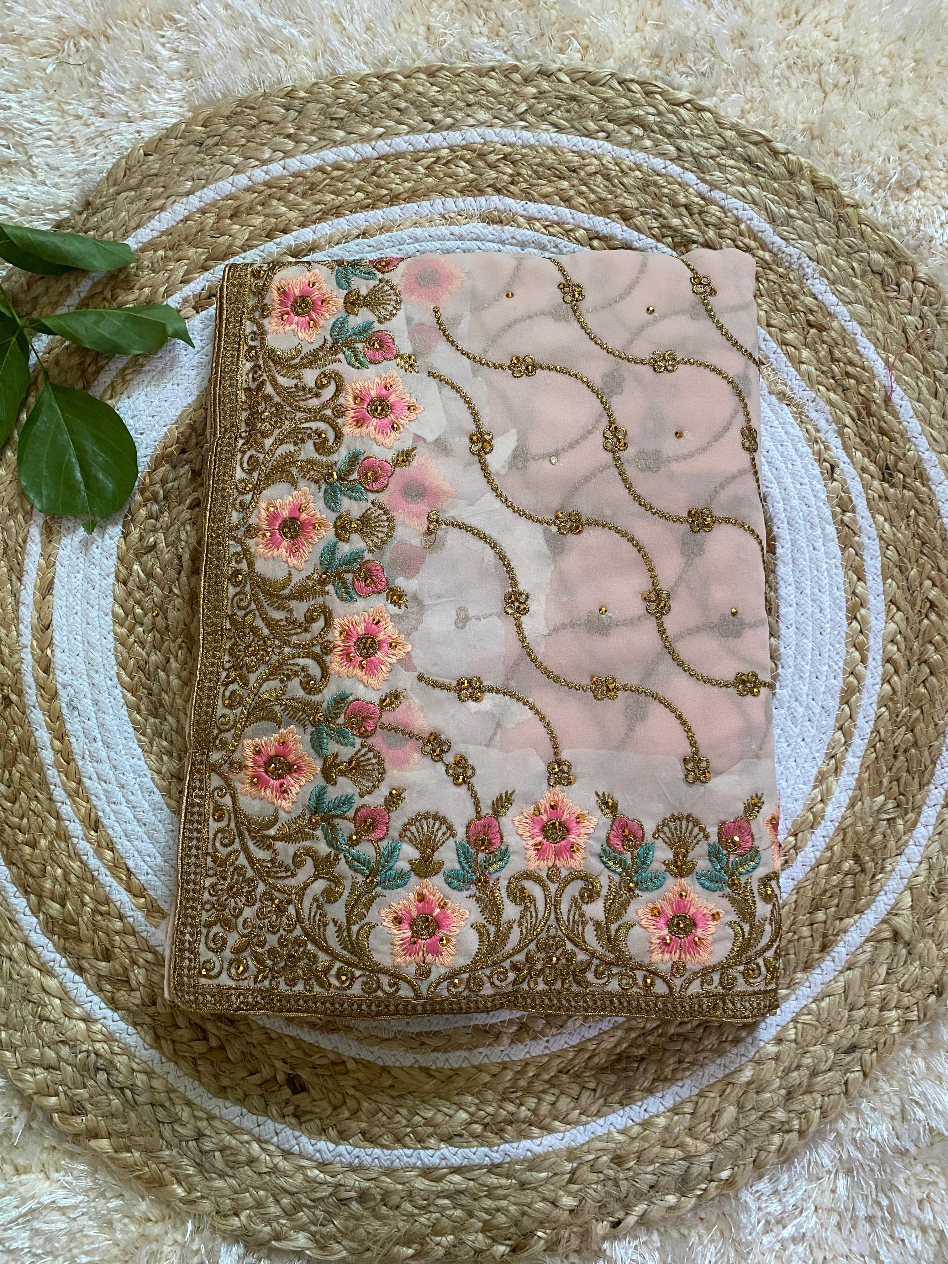 Folded pastel peach saree with intricate gold thread embroidery, featuring pink floral motifs and delicate vine patterns, beautifully arranged on a round woven jute mat with green leaves for styling. ✨