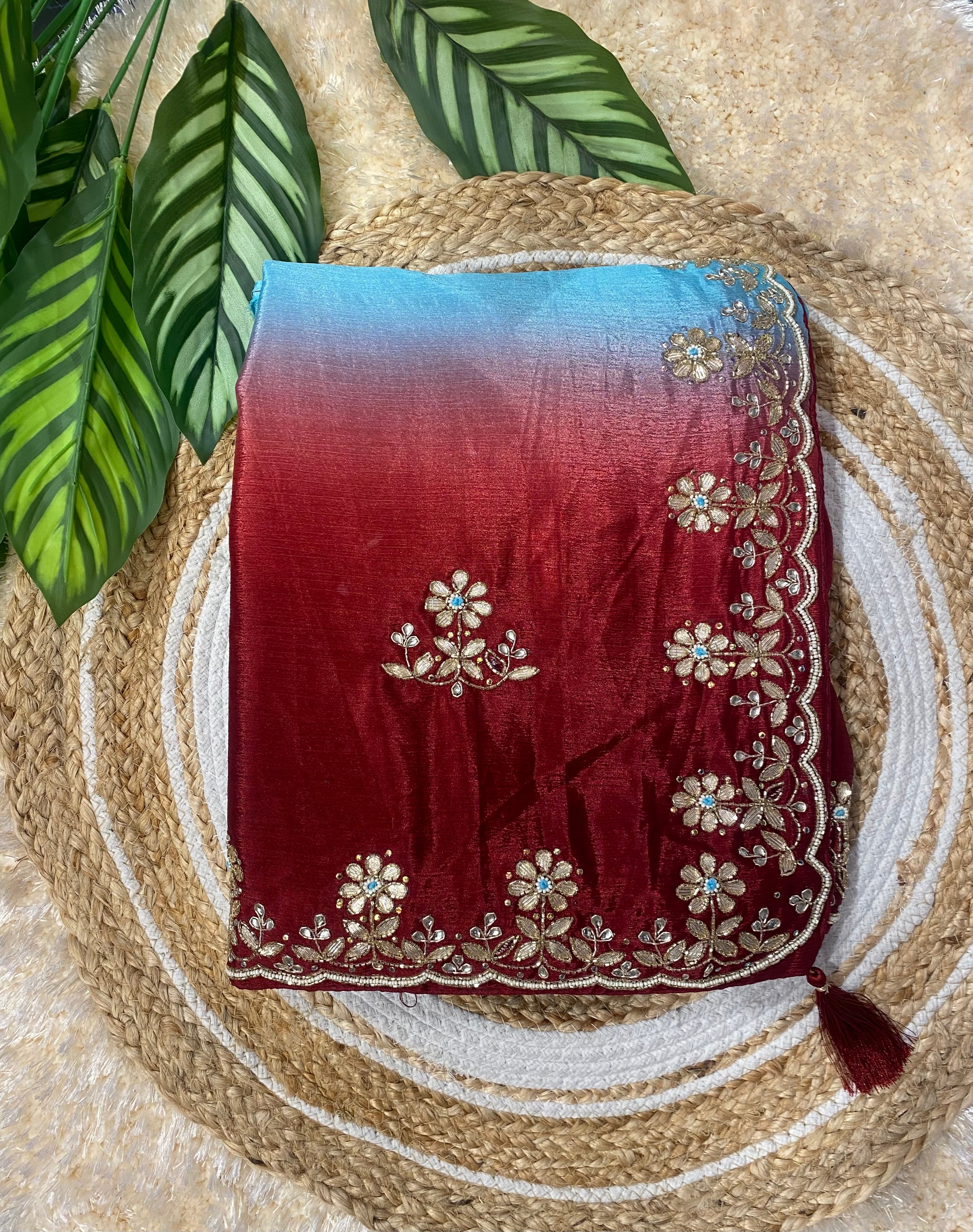 Folded saree displayed on a woven jute mat with green leaves in the background, featuring a rich red body with delicate gold floral embroidery and a gradient pallu transitioning from red to sky blue, finished with an ornate floral embroidered border and tassel detail. ✨