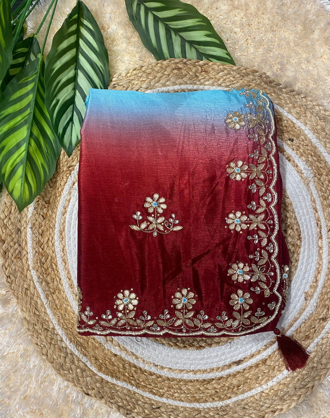 Folded saree displayed on a woven jute mat with green leaves in the background, featuring a rich red body with delicate gold floral embroidery and a gradient pallu transitioning from red to sky blue, finished with an ornate floral embroidered border and tassel detail. ✨
