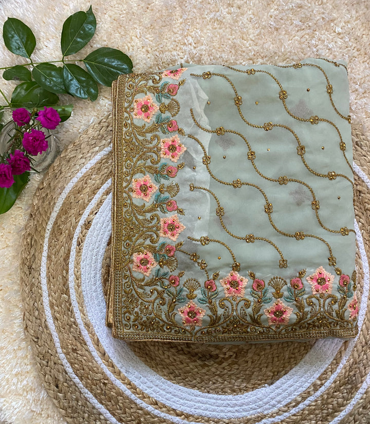 Folded sage green saree featuring intricate gold thread embroidery with pink floral motifs and teal leaf accents, paired with delicate vine patterns across the fabric, styled on a round woven jute mat with green leaves and a small vase of bright pink flowers. ✨