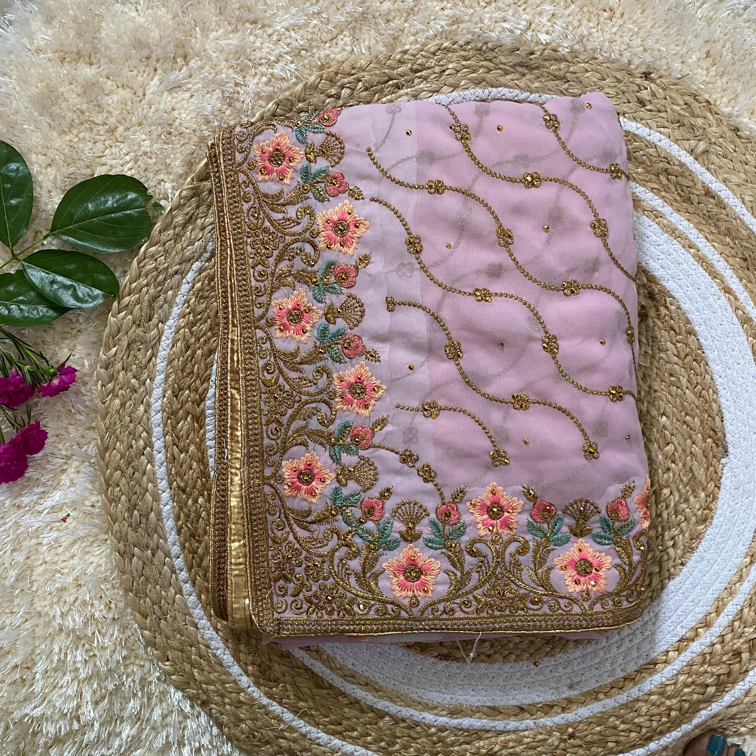 Folded blush pink saree featuring intricate gold thread embroidery with pink floral motifs and teal leaf accents, complemented by delicate vine patterns and a rich embroidered border, styled on a woven jute mat with greenery and flowers. ✨