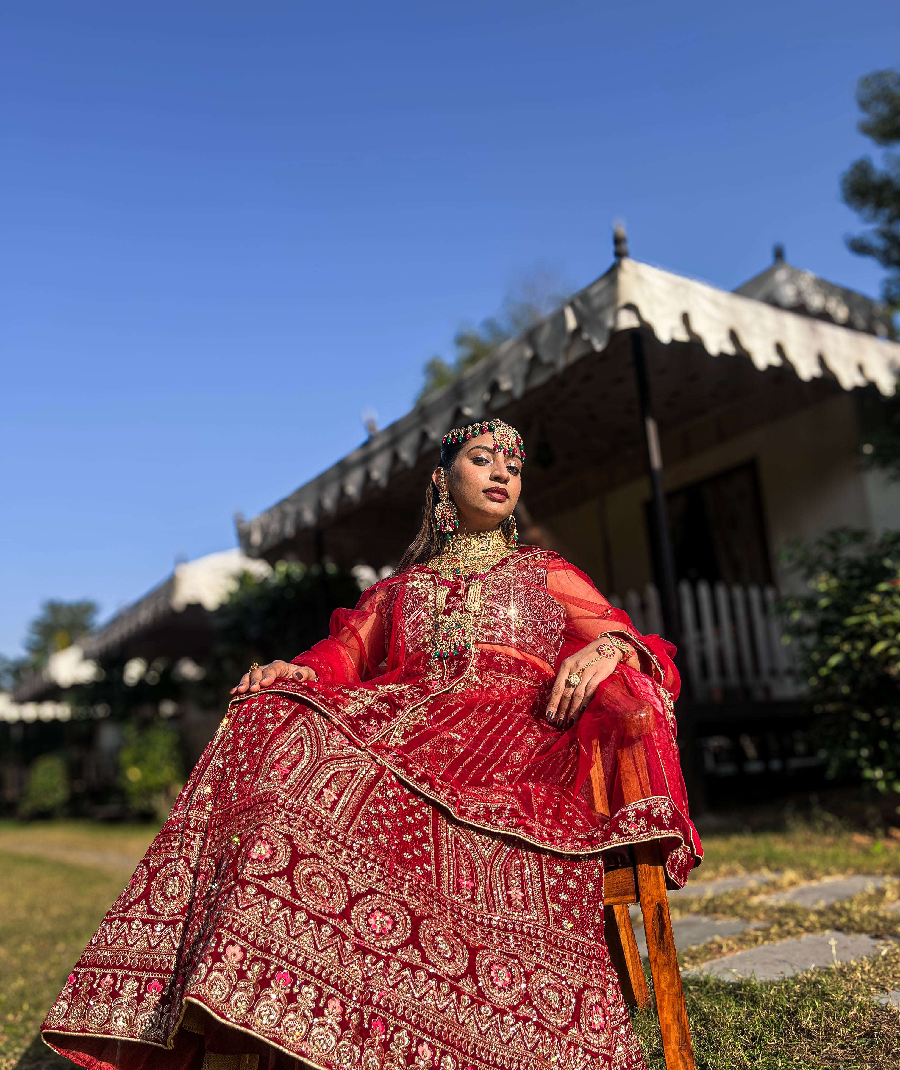 A bride sits outdoors in a richly embroidered red velvet bridal lehenga choli adorned with traditional gold jewelry, captured from a low angle.