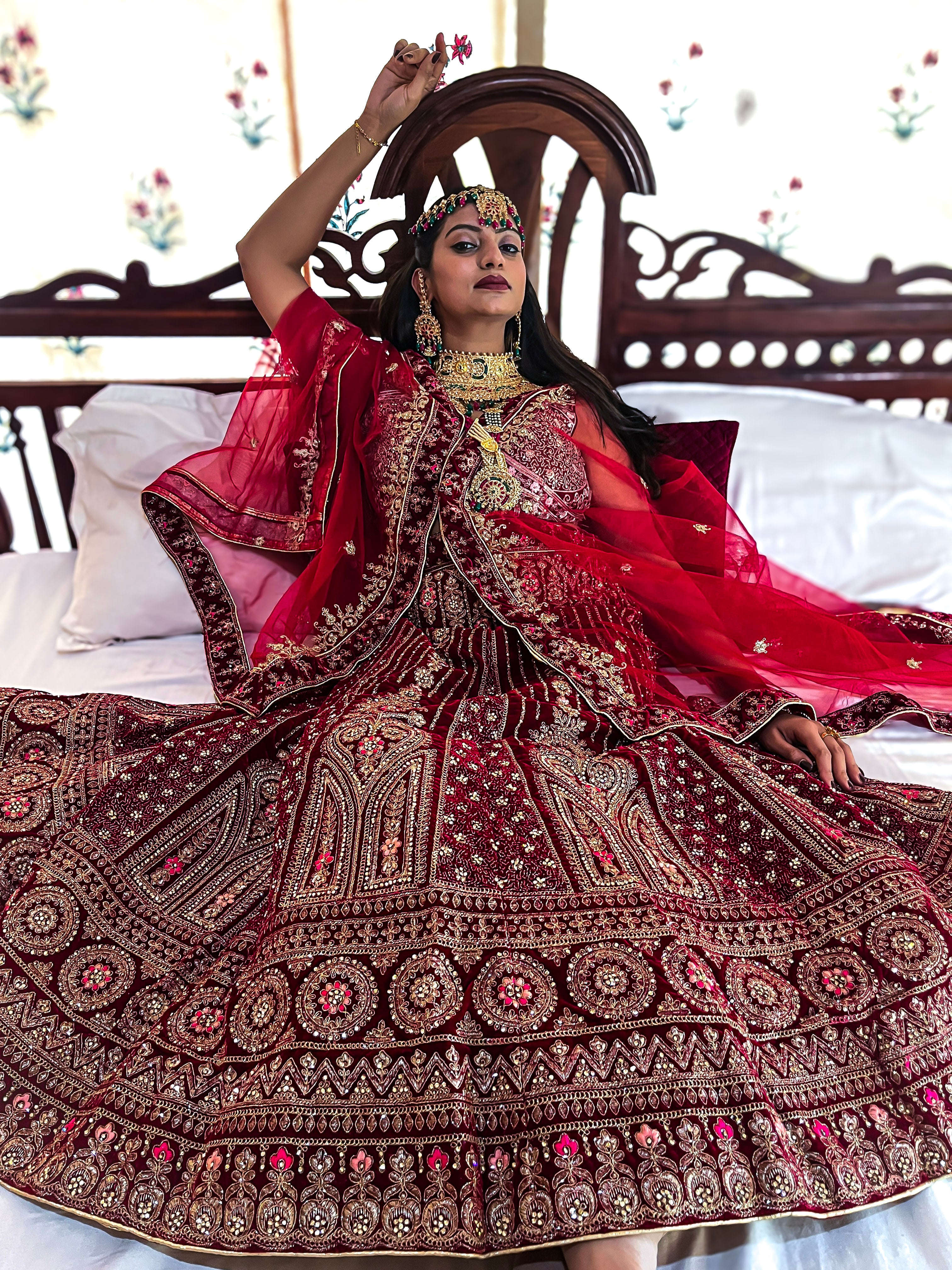 A bride poses on a wooden bed, showcasing her luxurious red velvet bridal lehenga choli with heavy gold zari embroidery and traditional jewelry.