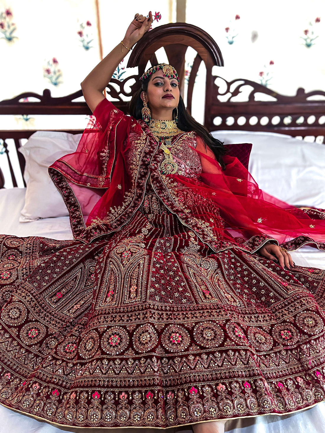 A bride poses on a wooden bed, showcasing her luxurious red velvet bridal lehenga choli with heavy gold zari embroidery and traditional jewelry.