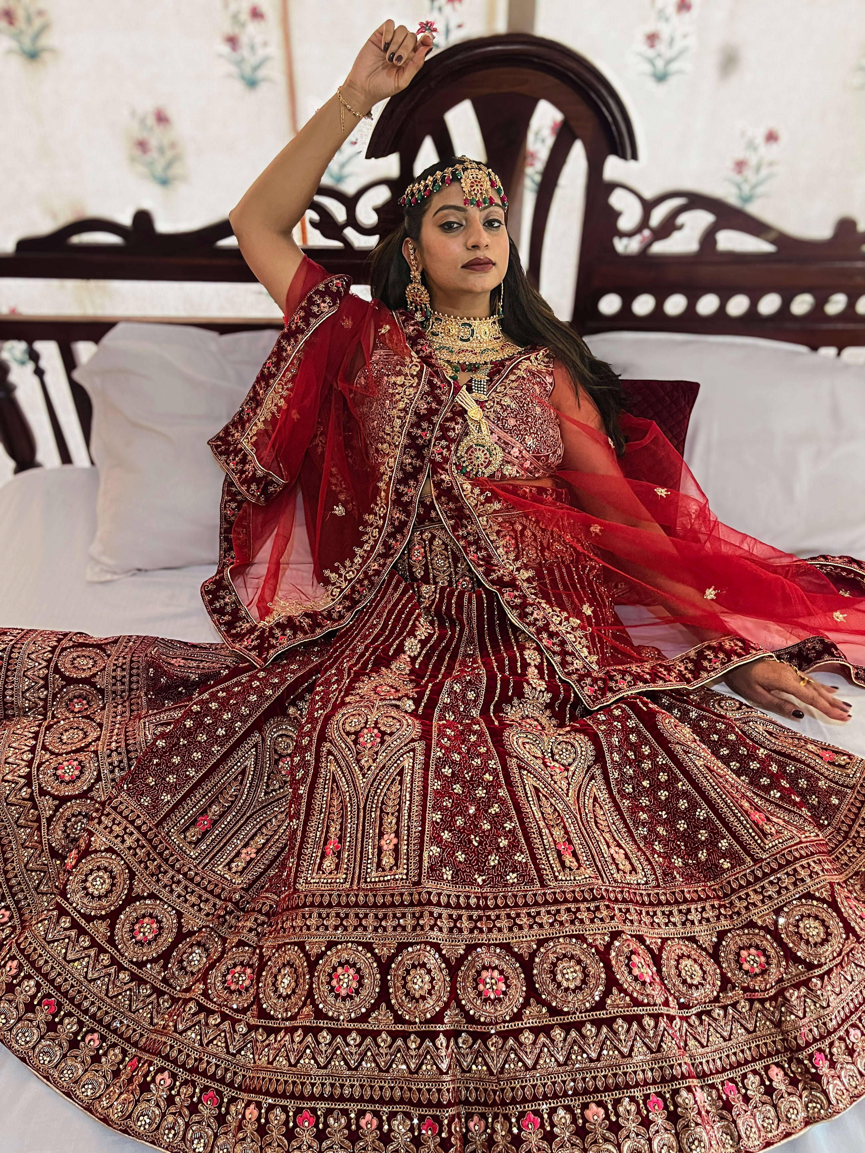 Portrait of an Indian bride seated in a royal setting, wearing a premium Red Velvet Lehenga Choli with heavy gold Zari handwork and traditional Kundan jewelry.