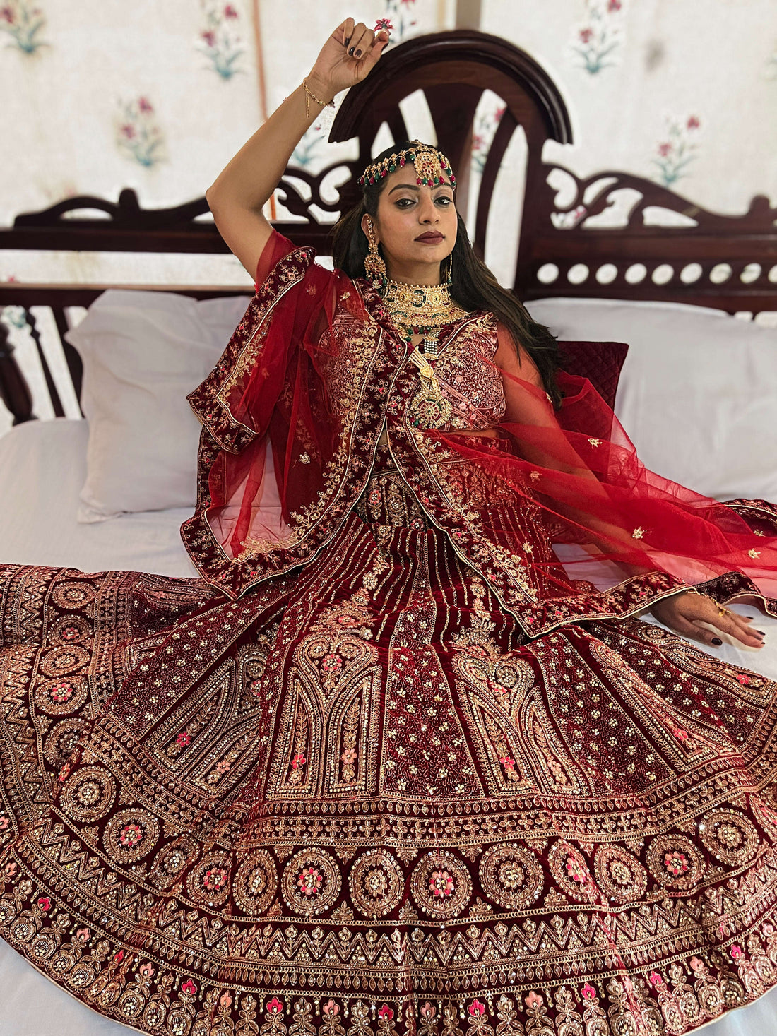 Portrait of an Indian bride seated in a royal setting, wearing a premium Red Velvet Lehenga Choli with heavy gold Zari handwork and traditional Kundan jewelry.