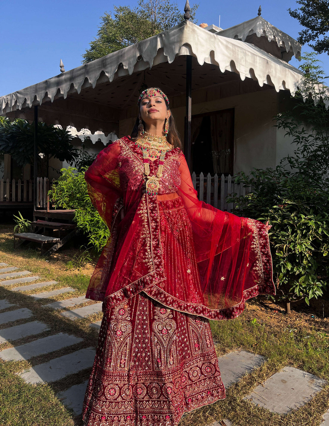 A bride poses outdoors in a heavily embroidered red velvet lehenga choli and sheer dupatta with gold zari work, showcasing traditional Indian bridal wear.