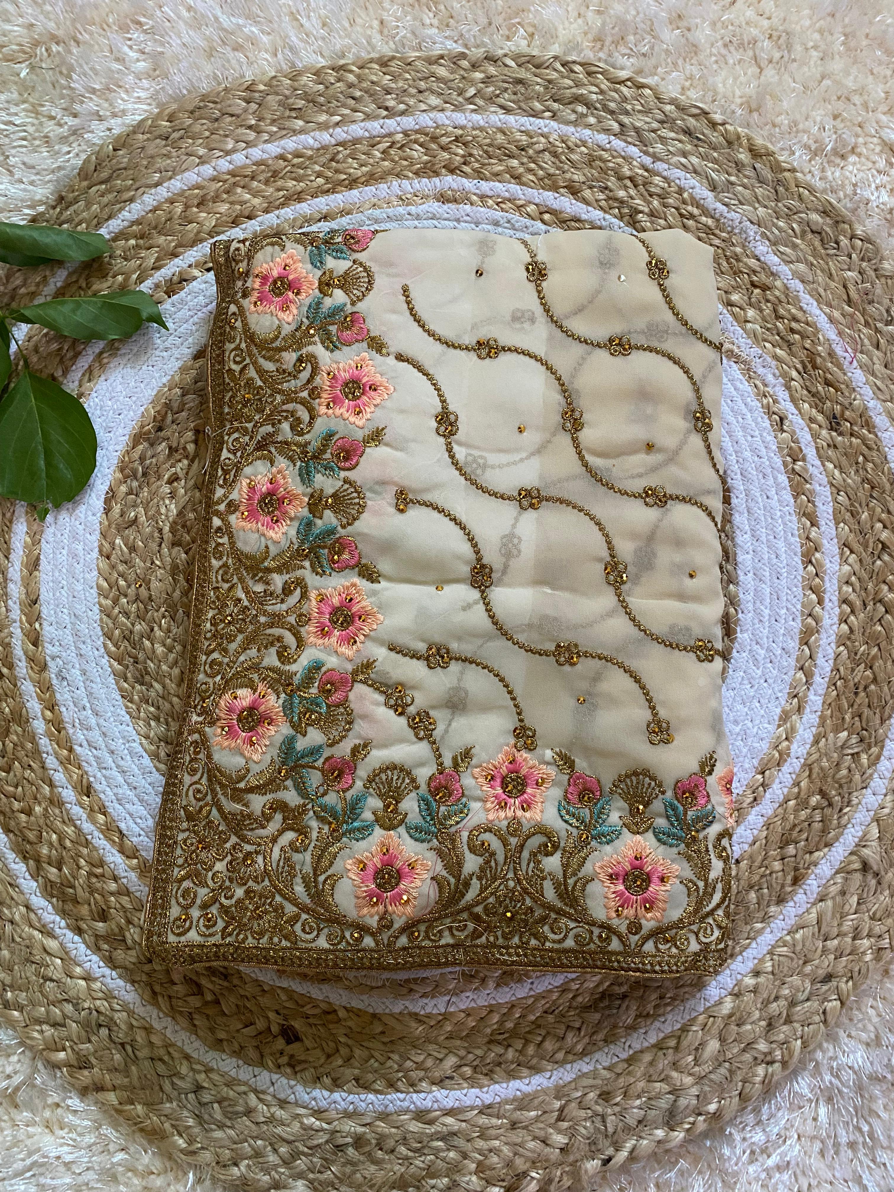 Folded cream-beige saree featuring intricate gold thread embroidery with pink floral motifs and teal leaf accents, paired with delicate vine patterns across the fabric, displayed on a round woven jute mat with green leaves for styling. ✨
