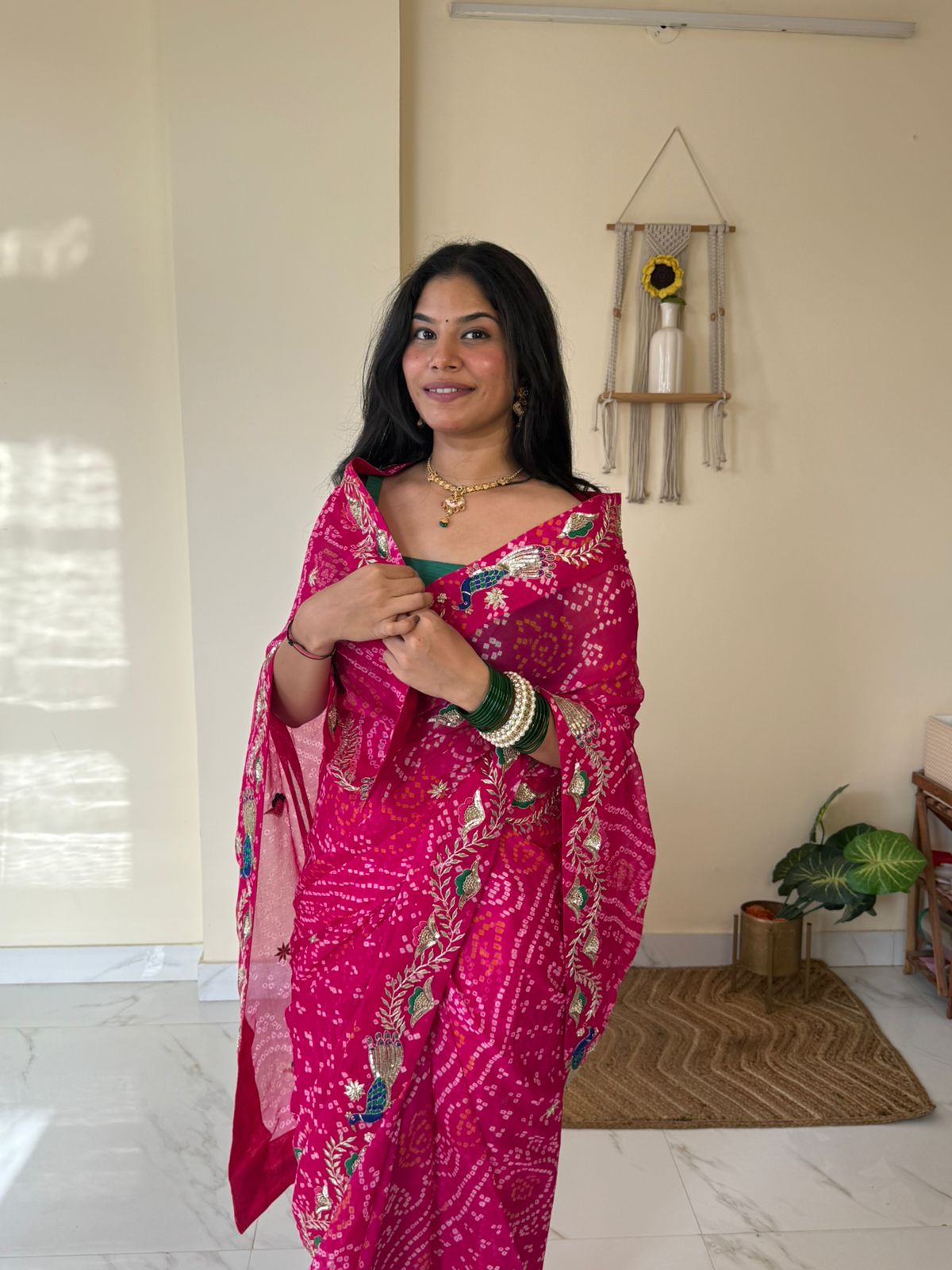 Close-up portrait of a woman draped in a vibrant rani-pink Bandhej saree featuring intricate peacock motifs, floral vine embroidery, and traditional handwork along the border. Styled with a green blouse, stacked bangles, and delicate gold jewelry, she poses gracefully in a bright indoor setting, showcasing the saree’s festive elegance and rich Rajasthani craftsmanship.