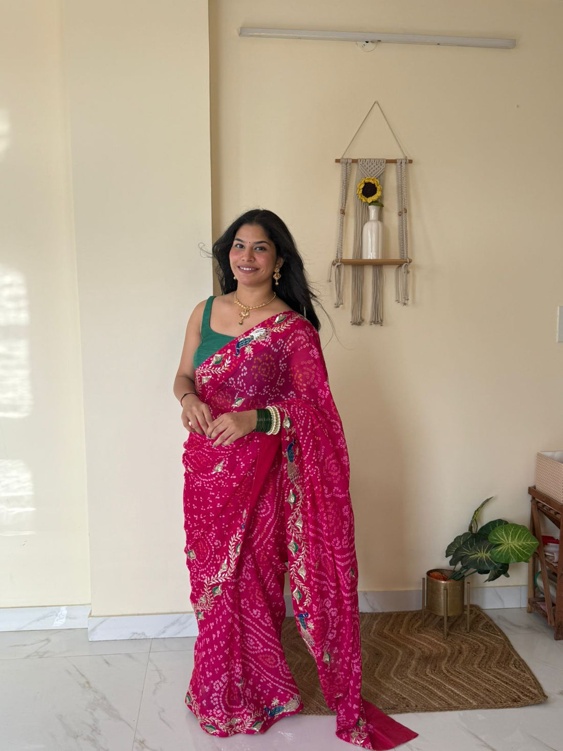 Woman wearing a vibrant rani-pink Bandhej saree adorned with intricate peacock and floral embroidery along the border, paired with a sleeveless green blouse and traditional jewelry. She stands gracefully in a bright, minimalist interior, showcasing the saree’s rich Rajasthani craftsmanship, festive charm, and elegant drape.