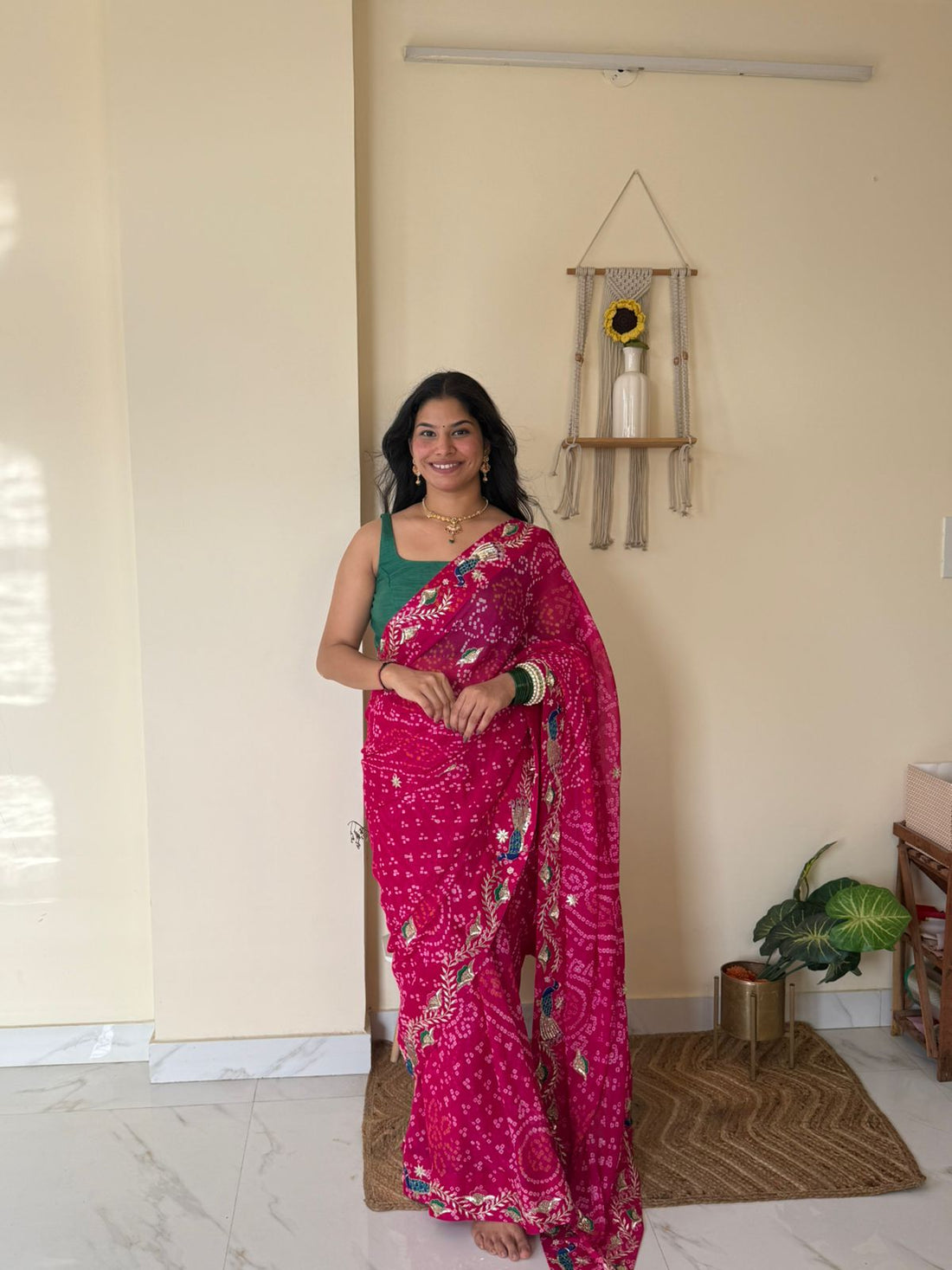 Full-length portrait of a woman wearing a vibrant rani-pink Bandhej saree embellished with intricate peacock motifs, floral vine embroidery, and traditional handwork borders. Paired with a contrasting green blouse, gold jewelry, and stacked bangles, she stands gracefully in a bright, minimalist indoor setting, showcasing the saree’s elegant drape, festive appeal, and rich Rajasthani heritage craftsmanship.