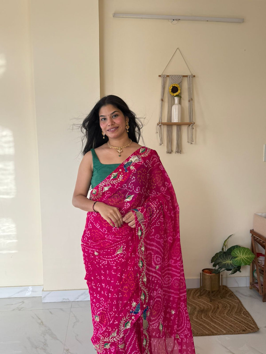Smiling woman draped in a vibrant rani-pink Bandhej saree featuring intricate hand-embroidered peacock motifs and floral border work, paired with a contrasting green blouse and traditional jewelry. Captured in a softly lit indoor setting that highlights the saree’s rich craftsmanship and festive elegance.