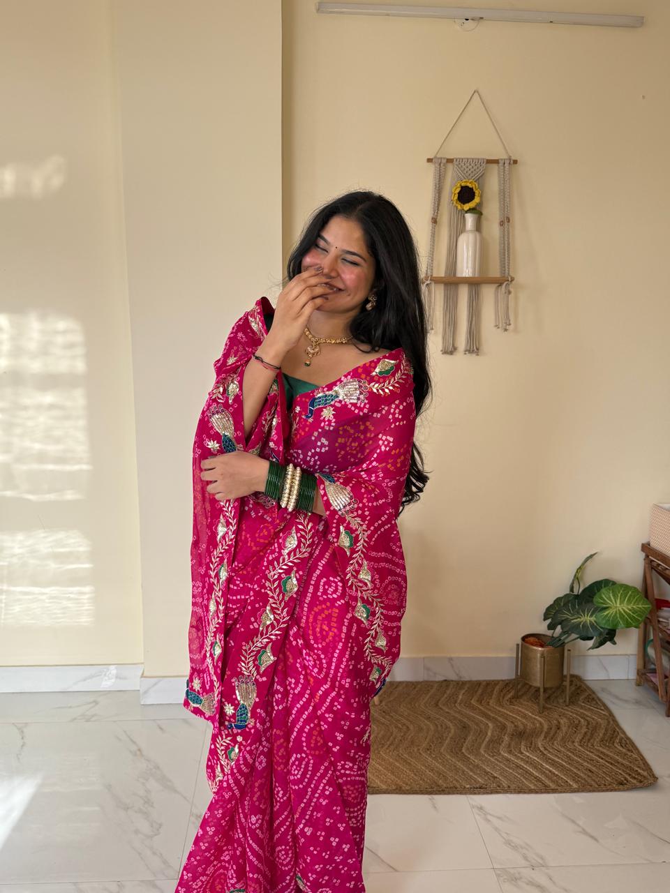 Woman draped in a vibrant rani-pink Bandhej saree featuring intricate peacock motifs, floral vine embroidery, and traditional handwork borders. Paired with a contrasting green blouse, stacked green-and-pearl bangles, and delicate gold jewelry, she is captured in a candid smiling moment in a bright indoor setting. The image highlights the saree’s graceful drape, festive elegance, and rich Rajasthani artisanal craftsmanship.
