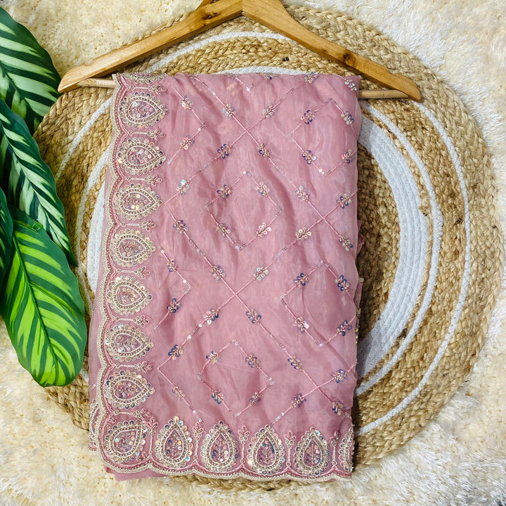 Folded blush pink saree displayed on a wooden hanger over a woven jute mat, featuring intricate paisley border embroidery with sequins and delicate diamond-pattern threadwork across the fabric. ✨